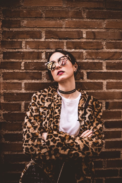 Stylish portrait of young serious model with crossed arms wearing trendy leopartd print faux fur coat, fashion sunglasses.