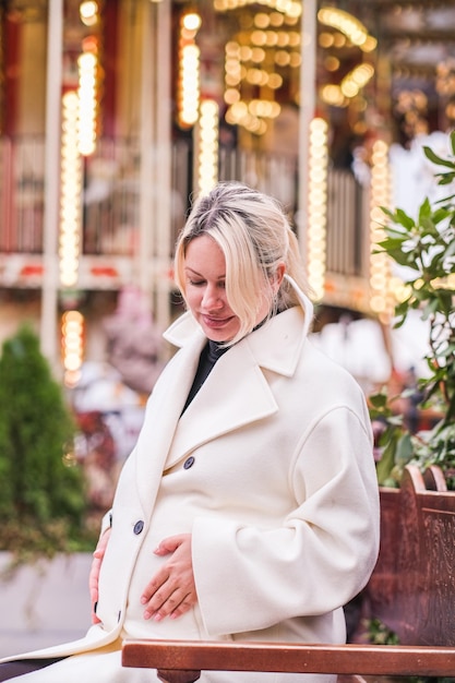 Stylish pregnant woman in a white dress posing on the background of carousels