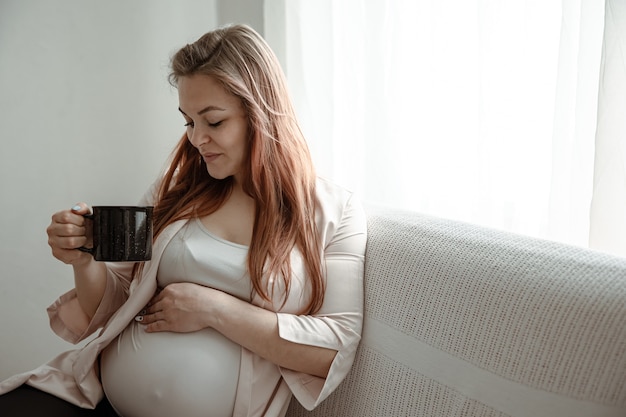 大きなお腹を持つスタイリッシュな妊婦が自宅のソファに座ってお茶やコーヒーを飲みます。