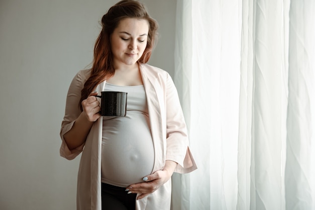 Stylish pregnant woman with a big belly with a cup of tea or coffee in her hands near the window.