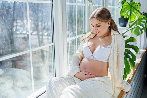 窓の近くの白いスーツを着たスタイリッシュな若い妊婦が優しい笑顔でお腹に触れ、出産を待っていますケアと母性愛と優しさ