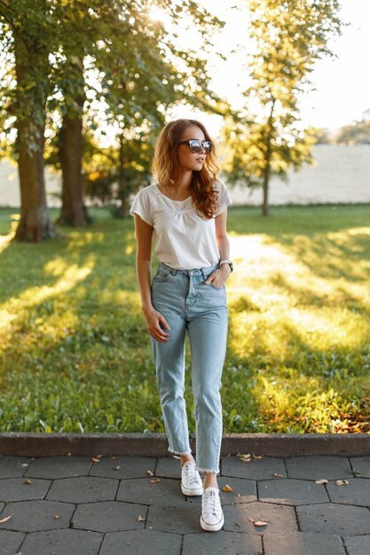 Stylish pretty hipster young woman in a white T-shirt in fashionable vintage jeans in trendy sunglasses in sneakers walks in the park among green trees.