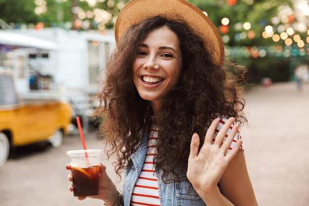 stylish pretty woman with curly brown hair wearing summer straw hat smiling and drinking cold tea from plastic cup outdoor, while waving hand