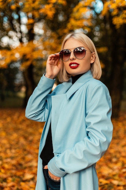 Elegante donna d'affari piuttosto giovane con un cappotto blu alla moda guarda attraverso gli occhiali da sole in un parco autunnale con fogliame autunnale giallo brillante