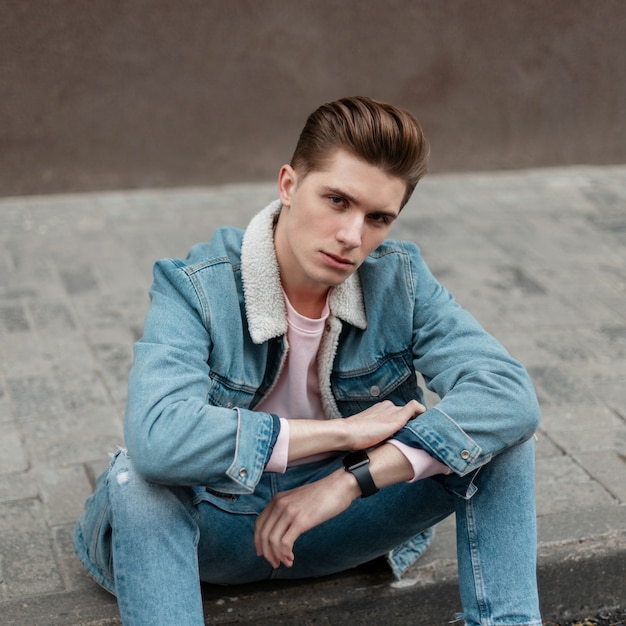 Stylish pretty young glamorous man in fashionable blue casual youth jeans clothes enjoys relaxing on tile near road on summer day in city. Handsome fashionable guy model. Street style. Vogue menswear.