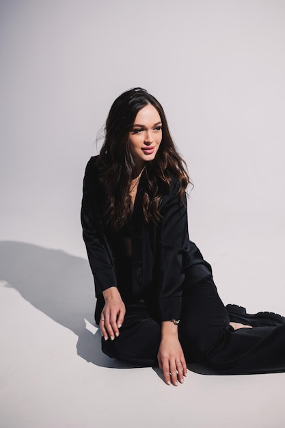 A stylish pretty young woman in a black classic suit is sitting on a white background A beautiful woman in a jacket and makeup on her face