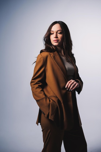 Stylish pretty young woman in brown classic suit sitting on gray background Beautiful woman in a jacket and with makeup on her face