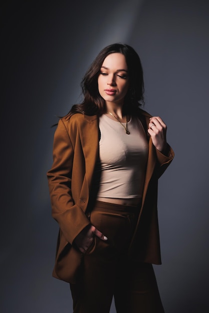 Stylish pretty young woman in brown classic suit sitting on gray background Beautiful woman in a jacket and with makeup on her face