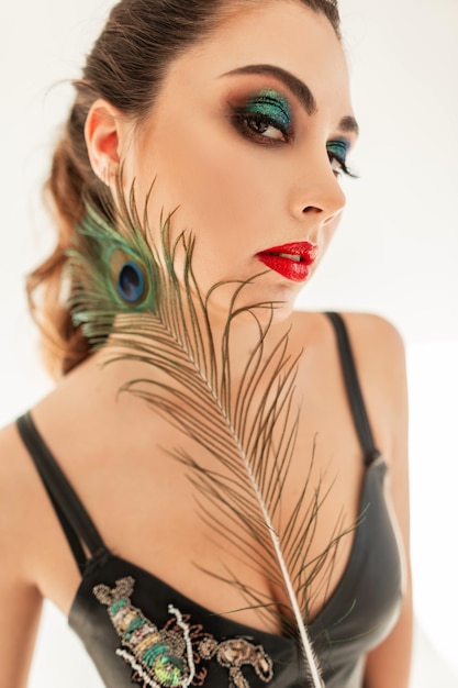 Stylish pretty young woman model with bright professional makeup, red lips and green shadows on the eyelids holding a colorful peacock feather in the studio