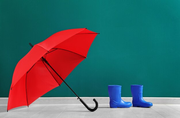 Stylish red umbrella and rubber boots near color wall