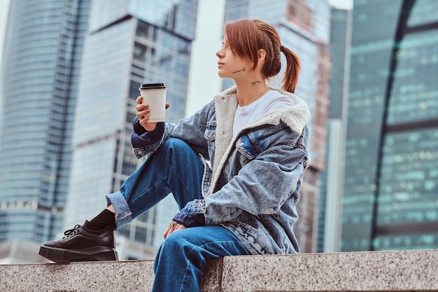 曇りの朝にモスクワ市の高層ビルの前に座って持ち帰り用のコーヒーを保持しているデニムのコートを着て彼女の顔にタトゥーを入れたスタイリッシュな赤毛の流行に敏感な女の子