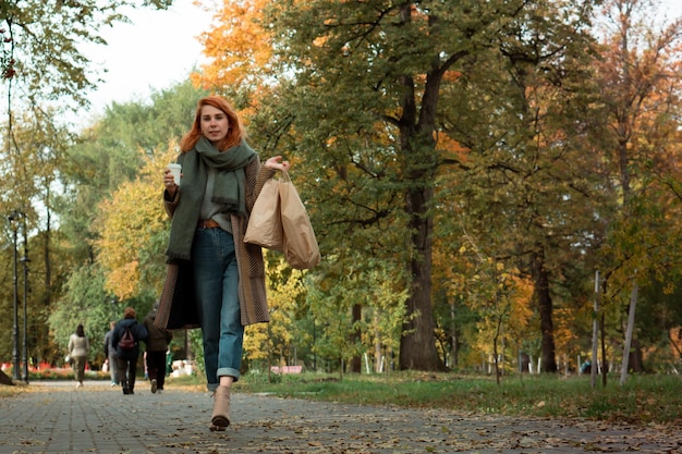 Фото Стильная рыжая молодая женщина в пальто с кофе и хозяйственными сумками гуляет в городском парке осенью