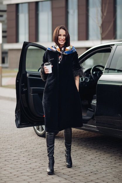 Stylish rich woman with cup of coffee next to her car.