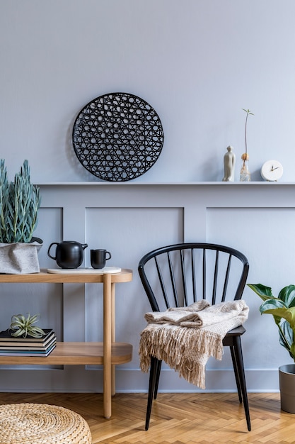 Foto interno elegante del soggiorno scandinavo con sedia nera di design, console in legno, piante, libri, decorazioni, teiera, pouf in rattan e accessori eleganti in un arredamento moderno.