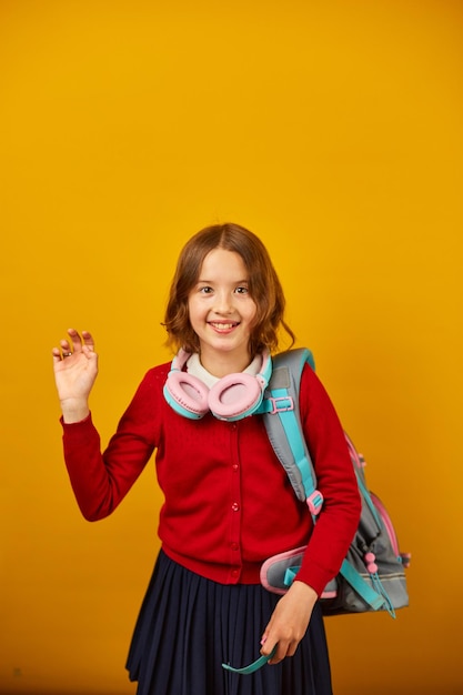 肩にバックパックを背負ったヘッドフォンをしたスタイリッシュな学校の10代の女の子が黄色のスタジオの背景に手を振って学校コンセプトのコピースペースに戻って友人に挨拶する