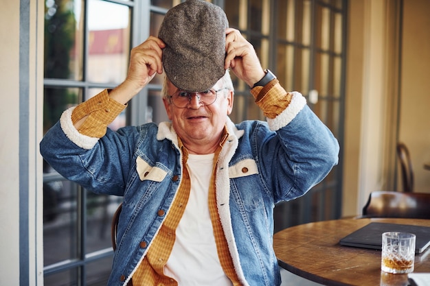 Stylish senior in fashionable clothes and in glasses sits in the cafe and holds hat.