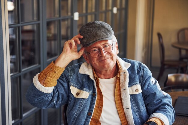 Stylish senior in fashionable clothes and in glasses sits in the cafe and holds hat.