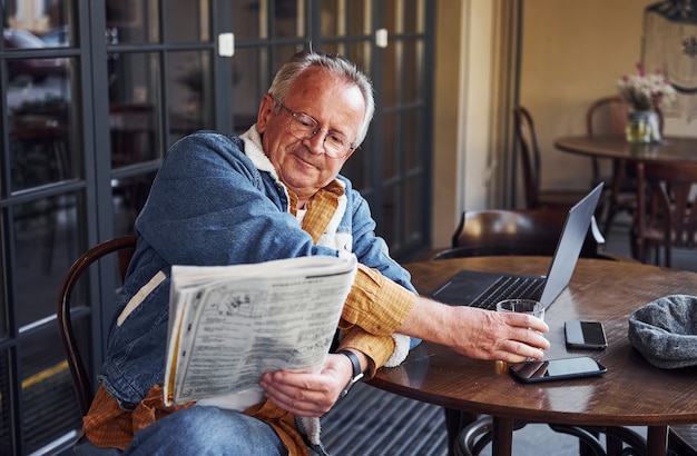 Stylish senior in fashionable clothes and in glasses sits in the cafe and reads newspaper.