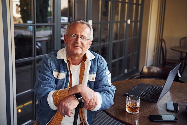 Stylish senior in fashionable clothes and in glasses sits in the cafe with drink and laptop on the table.