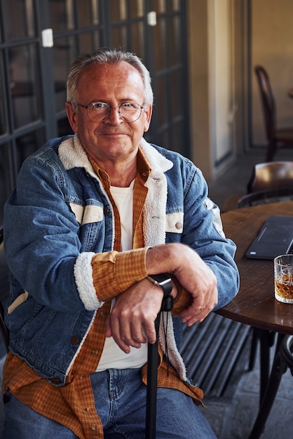 Stylish senior in fashionable clothes and in glasses sits in the cafe with drink and laptop on the table.