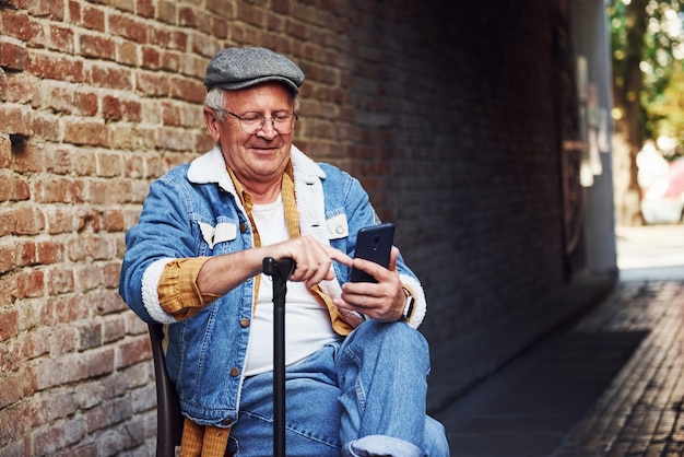 Stylish senior in fashionable clothes and in glasses sits on the chair and uses phone.