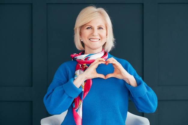 stylish senior woman. lady sitting on chair with heart shape hand sign. 