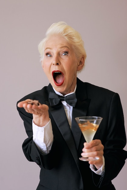 стильная старшая женщина в смокинге с бокалом с шампанским дует конфетти празднует новый год