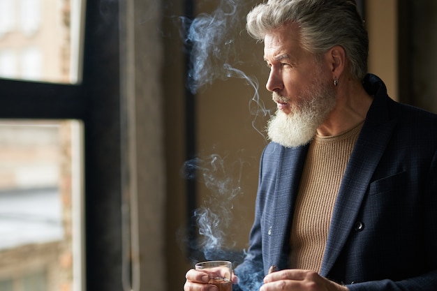 Stylish serious middle aged man with beard looking away while holding cigar and glass of