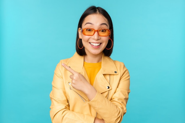 Stylish smiling asian woman in sunglasses pointing finger left showing advertisement banner standing in trendy yellow coat over blue background