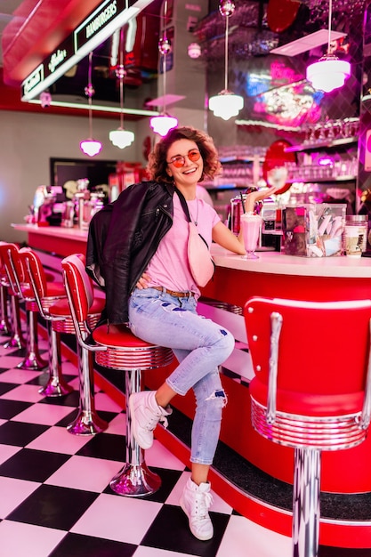 Stylish smiling beautiful woman in retro vintage 50's cafe sitting at bar drinking milk shake cocktail in pink t-shirt, balcj leather jacket and jeans, having fun in cheerful mood