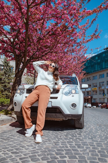 Stylish smiling beautiful woman standing near car blooming sakura tree on background