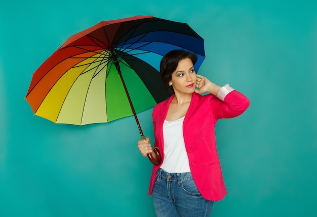 Stylish smiling girl in bright casual clothes under rainbow colored umbrella posing at azur studio background, copy space