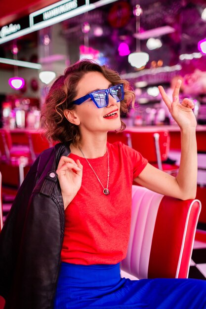 Stylish smiling woman in colorful outfit in retro vintage 50's cafe sitting at table