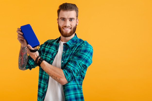 Stylish smiling young man holding wireless speaker listening to music on orange