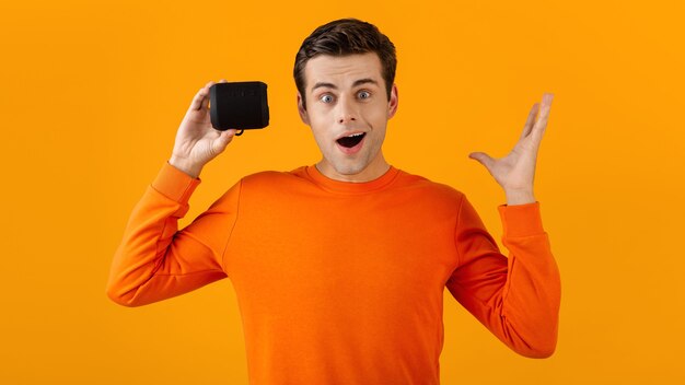 Stylish smiling young man in orange sweater holding wireless speaker happy listening to music having fun on orange