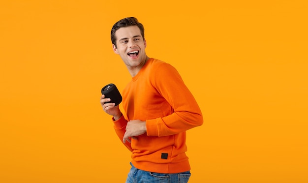 stylish smiling young man in orange sweater holding wireless speaker happy listening to music having fun on orange