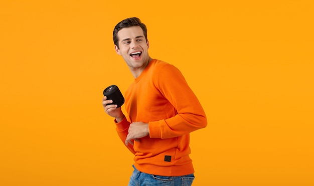 Stylish smiling young man in orange sweater holding wireless speaker happy listening to music having fun on orange