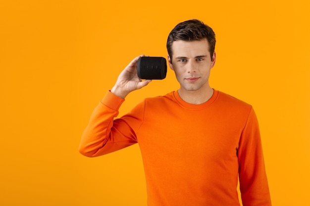 音楽を聞いて幸せなワイヤレススピーカーを保持しているオレンジ色のセーターでスタイリッシュな笑顔の若い男