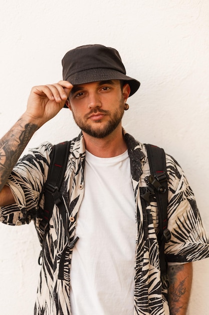 Photo stylish spanish fashionable man in fashion clothes with bucket hat and beach shirt loos at the camera near white wall on the street