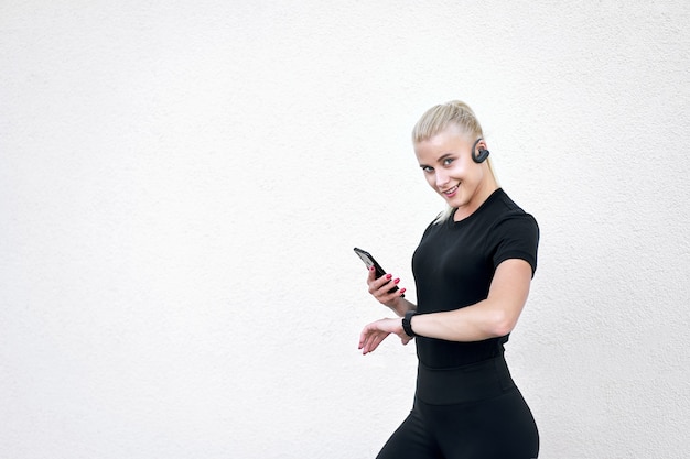Стильная спортивная женщина в черной спортивной одежде, слушает музыку и проверяет программу тренировок на умных часах