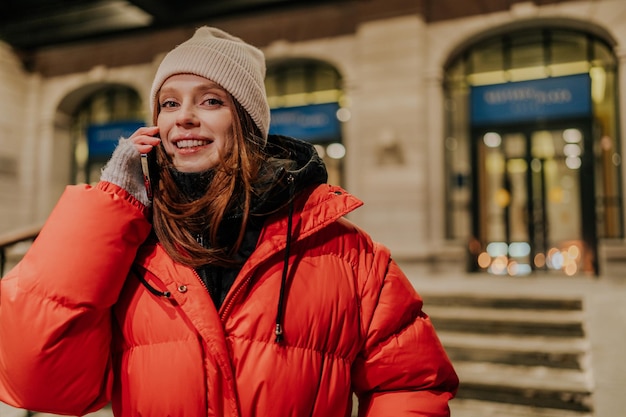 Stylish stedelijk portret van vrolijke roodharige jonge vrouw in warme winterkleding die praat met behulp van het vasthouden