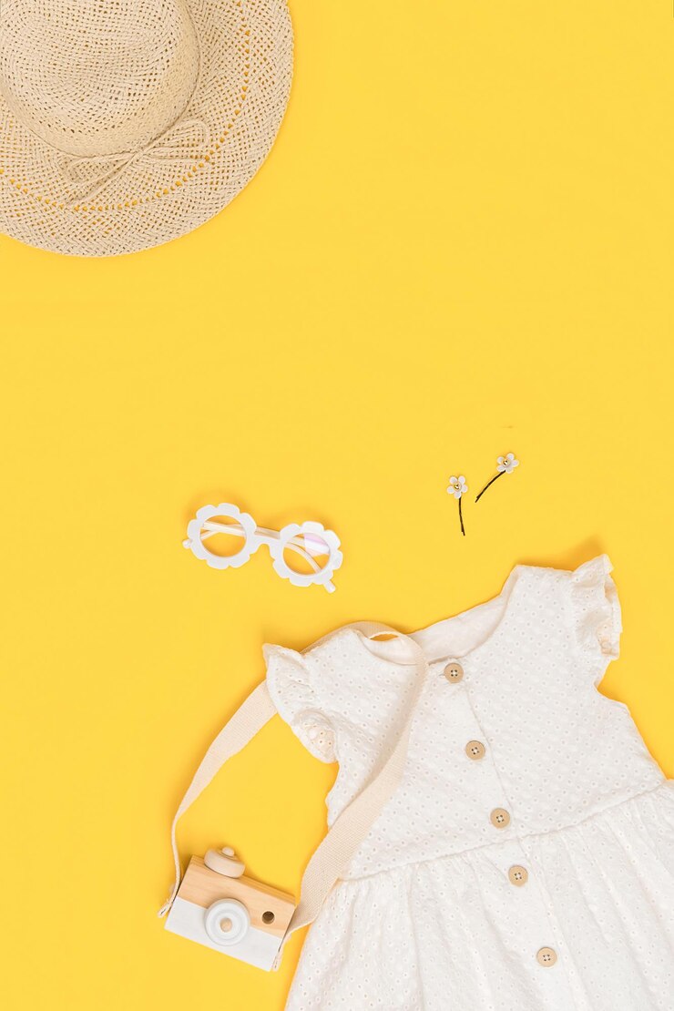 スタイリッシュな夏の子供服セット。白いドレス、麦わら帽子、サングラス、黄色の背景のアクセサリー。ファッションの女の子のルックブックのコンセプト。上面図コピースペースフラットレイ。