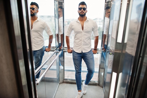 Stylish tall arabian man model in white shirt jeans and sunglasses posed at elevator inside