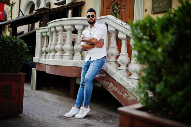 Elegante uomo arabo alto modello in camicia bianca jeans e occhiali da sole poste in strada della città barba attraente ragazzo arabo