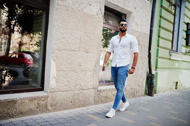 Stylish tall arabian man model in white shirt jeans and sunglasses posed at street of city Beard attractive arab guy