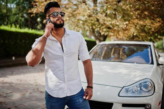 Stylish tall arabian man model in white shirt jeans and sunglasses posed at street of city Beard rich attractive arab guy against white suv car speaking on mobile phone