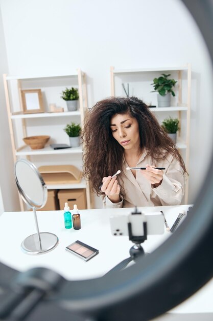 Stylish tanned handsome curly latin beauty blogger in linen\
shirt teach how to use small brush at home white interior copy\
space mockup banner influencer record video review blog using\
smartphone