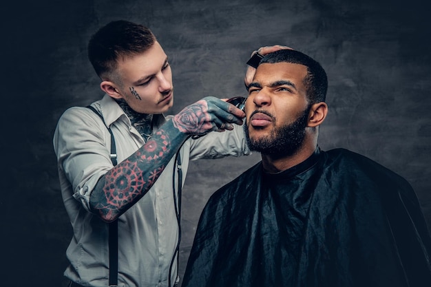 Il barbiere tatuato alla moda fa il taglio di capelli a un maschio con la barba nera.