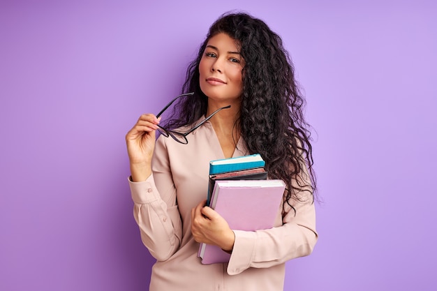 Stylish teacher in spectacles posing  holding books in hands isolated over purple wall