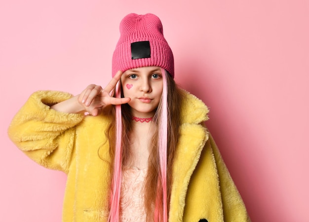 Stylish teenage girl with pink strands of hair in a pink hat yellow fluffy fur coat necklace and a painted heart on her face shows victoria with her fingers